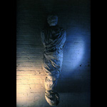 body wrapped in white cloth mounted on brick wall between two windows lit by two lights out of frame